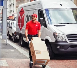 A man pushing a cart of boxes for delivery with his delivery van behind him.